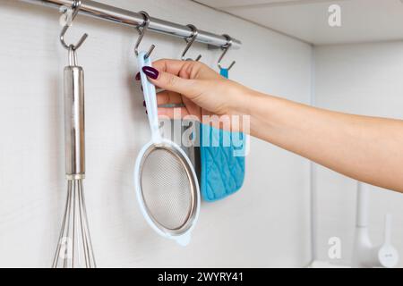 la femme au foyer retire la crépine de cuisine du crochet sur le rail. rail de cuisine avec ustensiles. tamis métallique de cuisine. Photo de haute qualité Banque D'Images