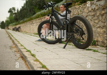 Vélo électrique, moto, vélo à partager. E-bike garé dans la ville, contre un bel homme relaxant. Service de partage de vélos en ville. Durabilité. Banque D'Images