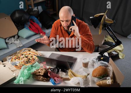 Homme barbu slatternly assis devant un ordinateur portable et tenant un smartphone parmi les restes, la nourriture, les sacs et les ordures sur fond Banque D'Images