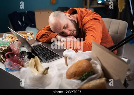 Homme millénaire épuisé dormant sur un bureau près d'un ordinateur portable, travaillant dans une pièce en désordre, fatigué de surmenage. Bourreau de travail souffrant de fatigue chronique à Banque D'Images