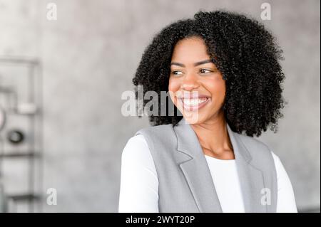 Une femme d'affaires afro-américaine rayonnante sourit avec confiance, debout dans un environnement de bureau moderne. Sa tenue professionnelle et son comportement joyeux projettent à la fois l'accessibilité et l'autorité. Banque D'Images
