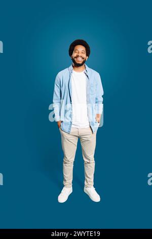 Un homme joyeux avec une coiffure afro naturelle se tient confiant, les mains dans les poches, dans une chemise en denim décontractée, et un sourire authentique, sur fond bleu profond, pleine longueur Banque D'Images