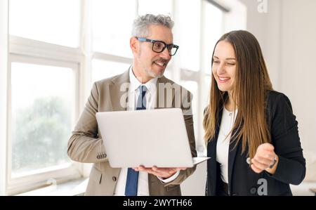 Homme d'affaires senior avec les cheveux gris partage un moment léger avec une collègue tout en travaillant sur un ordinateur portable, leur camaraderie facile évidente dans un bureau moderne dans une atmosphère de travail positive Banque D'Images