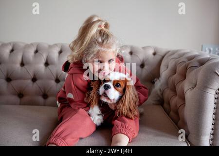 Belle fille blonde enfant est assis à la maison sur un canapé, jouant avec et embrassant le chien, créant des moments de joie et de compagnie avec l'animal de compagnie. Banque D'Images