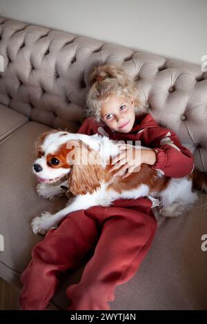 Belle fille blonde enfant est assis à la maison sur un canapé, jouant avec et embrassant le chien, créant des moments de joie et de compagnie avec l'animal de compagnie. Banque D'Images