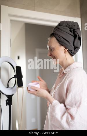 Blogueur souriant et montrant le pot de crème cosmétique dans la salle de bain devant le téléphone Banque D'Images