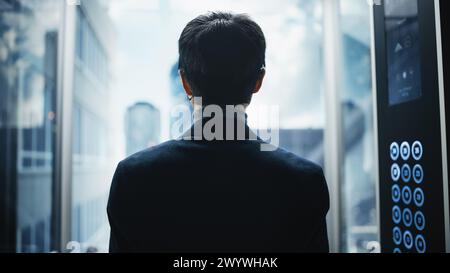 Élégant homme d'affaires japonais chevauchant l'ascenseur en verre au bureau dans le centre d'affaires moderne. Homme regardant les gratte-ciel modernes du centre-ville par la fenêtre Panorama dans l'ascenseur. Retour tourné vers appareil photo. Banque D'Images