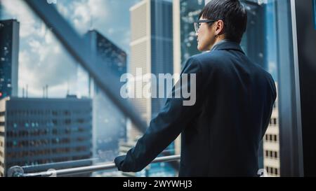 Élégant homme d'affaires japonais chevauchant l'ascenseur en verre au bureau dans le centre d'affaires moderne. Homme regardant les gratte-ciel modernes du centre-ville par la fenêtre Panorama dans l'ascenseur. Retour tourné vers appareil photo. Banque D'Images