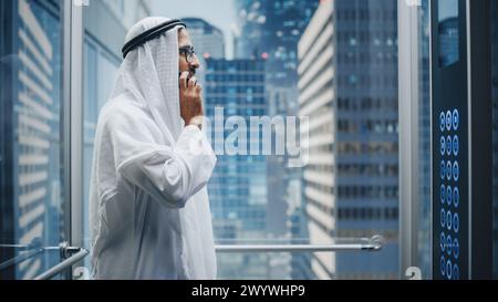 Homme d'affaires musulman prospère dans l'ascenseur en verre blanc traditionnel de Kandura au bureau dans le centre d'affaires moderne. Homme parlant sur un téléphone dans un ascenseur. Saoudien, émirati, concept d'homme d'affaires arabe. Banque D'Images