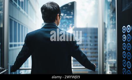 Élégant homme d'affaires japonais chevauchant l'ascenseur en verre au bureau dans le centre d'affaires moderne. Homme regardant les gratte-ciel modernes du centre-ville par la fenêtre Panorama dans l'ascenseur. Retour tourné vers appareil photo. Banque D'Images