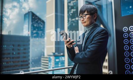 Homme d'affaires japonais élégant réussi chevauchant l'ascenseur en verre au bureau dans le centre d'affaires moderne. Beau sourire d'homme tout en utilisant smartphone, écrire un message texte, vérifier les médias sociaux dans un ascenseur. Banque D'Images