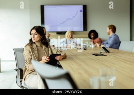 Femme d'affaires concentrée avec un bloc-notes réfléchit lors d'une réunion d'équipe, avec des collègues engagés dans la discussion en arrière-plan Banque D'Images
