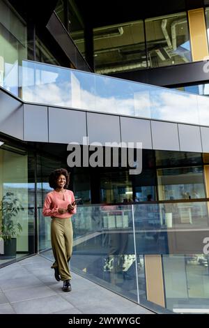 Femme d'affaires afro-américaine avec un sourire radieux marche vigoureusement près d'un immeuble de bureaux, exsudant la confiance et l'élégance dans sa tenue élégante Banque D'Images