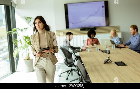 Leader féminin déterminé se tient au premier plan avec une tablette, s'adressant à son équipe attentive lors d'une réunion. Banque D'Images