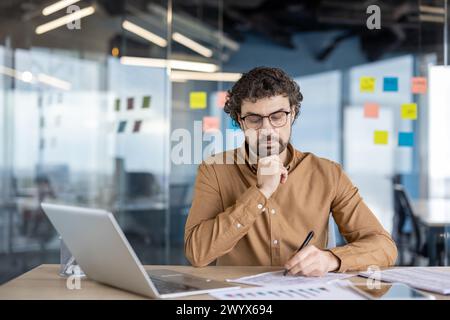 Homme d'affaires sérieux pensant derrière le travail de papier à l'intérieur du bureau, l'homme remplit des formulaires, des rapports et des factures. Un employé utilise un ordinateur portable pour la paperasserie. Banque D'Images