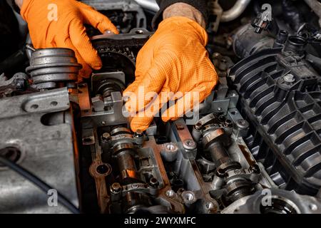 Vue détaillée des mains d'un mécanicien automobile, protégées par des gants orange, alors qu'ils travaillent attentivement à la réparation d'un moteur de voiture ouvert dans un service automobile Banque D'Images