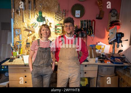 Heureux couple de menuisiers portant des combinaisons en denim debout dans une pièce artisanale ensemble près de la table des outils Banque D'Images