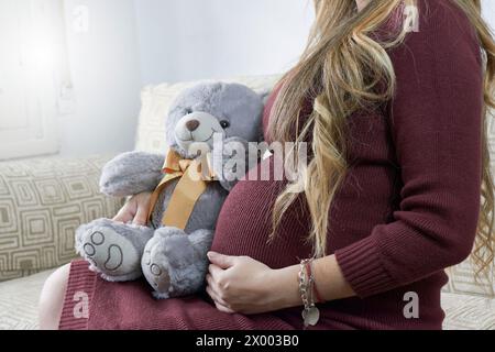 Une femme aux cheveux blonds et une robe rouge est assise sur un canapé. Elle est enceinte, à côté d'elle se trouve un ours en peluche gris et blanc avec un arc orange. Banque D'Images