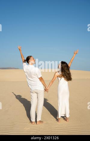 Vue arrière de jeunes voyageurs de couple pieds nus se regardant debout sur un terrain sablonneux avec les mains levées et avec des ombres contre les nuages Banque D'Images