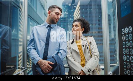 Deux collègues de bureau se regardant tout en roulant dans Glass Elevator dans un centre d'affaires moderne. Spécialiste caucasien masculin et responsable féminine noire d'Amérique latine discutez en toute décontraction dans l'ascenseur. Banque D'Images