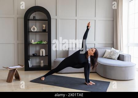 Une femme pratiquant le yoga à la maison, effectuant l'exercice Utthita Parshvakonasana, une pose avec un angle latéral étendu, la formation dans un sport noir overa Banque D'Images