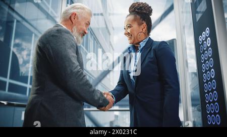 Homme d'affaires d'âge moyen parlant avec un partenaire d'investissement international tout en montant l'ascenseur de verre au bureau dans un centre d'affaires moderne. Les collaborateurs d'entreprise se mettent à la main sur un accord réussi. Banque D'Images