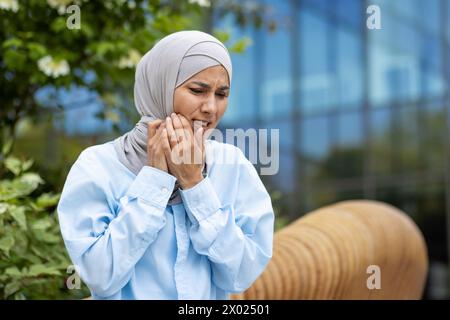 Portrait de femme portant le hijab et la chemise bleu clair tout en touchant la joue avec les deux mains sur le fond de l'extérieur du bâtiment vitreux. Femme plissée présentant un symptôme de sensibilité dentaire. Banque D'Images
