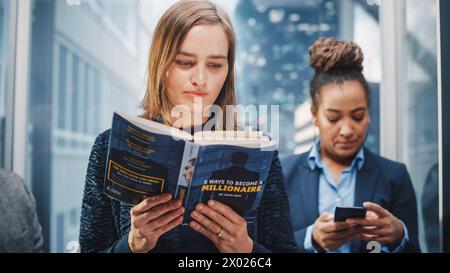 Confiant jeune femme Riding Glass ascenseur au bureau dans le centre d'affaires moderne. Manager junior lisant un livre financier intitulé « 5 façons de devenir millionnaire » dans un ascenseur. Banque D'Images