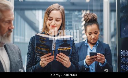 Confiant jeune femme Riding Glass ascenseur au bureau dans le centre d'affaires moderne. Manager junior lisant un livre financier intitulé « 5 façons de devenir millionnaire » dans un ascenseur. Banque D'Images