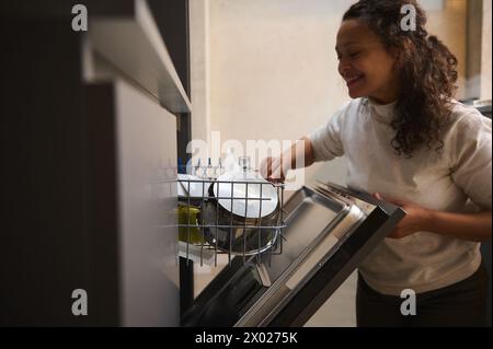 Portrait authentique d'une brune multi ethnique louée frisée, d'une femme au foyer, d'une femme souriante heureuse utilisant un lave-vaisselle moderne dans la cuisine. Entretien ménager. Banque D'Images