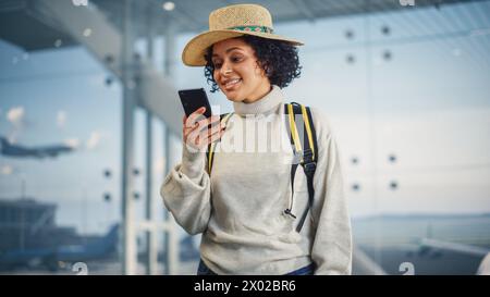 Terminal de l'aéroport : Happy Travelling Black Woman regarde autour de la recherche des portes de vol et de l'avion, utilise smartphone, vérification de la destination du voyage sur Internet. African American Female se demandant dans Airline Hub Banque D'Images