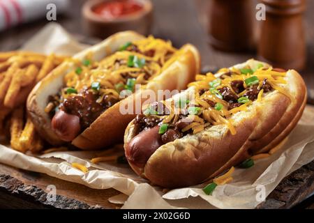 Deux hot-dogs au Chili avec fromage cheddar râpé, oignons verts hachés et frites assaisonnées sur un plateau en bois Banque D'Images