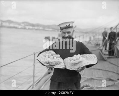 LE HMS HERMIONE EN PATROUILLE DANS L'ATLANTIQUE, ESCORTANT LE SOUS-MARIN HMS REGENT ENDOMMAGÉ PAR LES INTEMPÉRIES. 24 JANVIER 1942, À BORD DU CROISEUR HMS HERMIONE. LE SOUS-MARIN A ÉTÉ EMMENÉ À PONTA DELGADO, UN PORT NEUTRE DES AÇORES PORTUGAISES, POUR 24 HEURES DE RÉPARATIONS. - Un marin britannique avec une armure d'œufs achetés à des commerçants portugais qui est venu aux côtés du HMS HERMIONE dans des bateaux à rames Banque D'Images