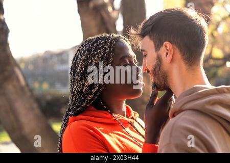 Une jeune femme africaine avec des cheveux tressés touche doucement le visage d'un homme - regards enfermés dans une tendresse étreinte. Amants multiraciaux au coucher du soleil - diverse lo Banque D'Images