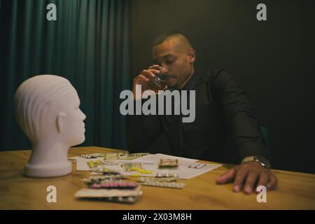 Homme afro-américain buvant du whisky alcoolisé dans un casino. Il y a de l'argent dispersé sur la table, pilules tête de mannequin. Banque D'Images