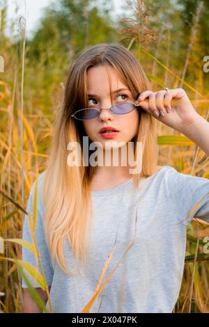 Belle jeune fille portant un t-shirt gris vierge et un Jean noir dans des lunettes bleues posant contre l'herbe haute vert et jaune au début de l'automne chaud. Banque D'Images