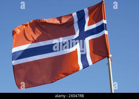 Un drapeau rouge vif avec une croix bleue encadrée en blanc, représentant la Norvège, flotte dans la brise sous un ciel sans nuages. Banque D'Images