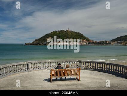 Un couple de personnes âgées se repose assis sur un banc tout en profitant du paysage devant eux, une belle île avec une grande sculpture sur le dessus Banque D'Images