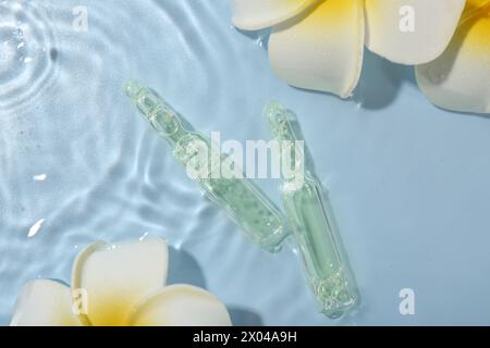 Ampoules de soins de la peau et belles fleurs de plumeria dans l'eau sur fond bleu clair, pose à plat Banque D'Images