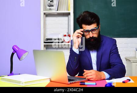 Professeur barbu sérieux dans des lunettes assis au bureau travaillant avec un ordinateur portable dans la salle de classe. Étudiant masculin étudiant en ligne. E-learning au collège universitaire ou Banque D'Images