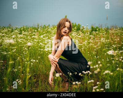 Belle jeune fille dans une robe de soirée noire pose accroupie devant un champ de marguerites sur une journée d'été nuageuse. Portrait d'un mannequin féminin à l'extérieur. Banque D'Images