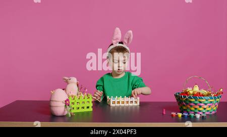 Heureux tout-petit avec des oreilles de lapin arrangeant panier rempli d'œufs peints, créant des arrangements festifs pour la célébration du dimanche de pâques. Joyeux petit enfant aime l'art et l'artisanat. Caméra B. Banque D'Images