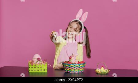 Jeune fille excitée arrangeant des œufs peints dans un panier pour préparer la célébration des vacances de pâques, créant des arrangements festifs. Enfant heureux ludique avec des oreilles de lapin, activité créative. Caméra A. Banque D'Images