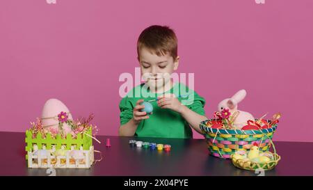 Petit garçon joyeux fabriquant des décorations de pâques faites à la main en peignant avec de l'aquarelle et des pinceaux. Petit jeune enfant utilisant l'art fournit des oeufs de coloration pour les vacances, activité de décoration. Caméra A. Banque D'Images