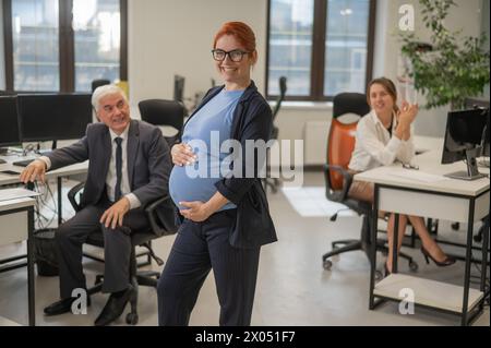 Une femme enceinte heureuse se tient au milieu du bureau à côté d'une femme caucasienne et d'un homme âgé travaillant sur des ordinateurs. Banque D'Images