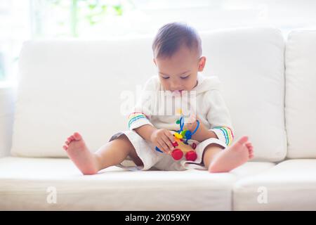 Bébé asiatique jouant avec un train de jouets coloré dans une chambre blanche ensoleillée. Bébé avec des jouets éducatifs. Enfant assis sur un canapé blanc. Banque D'Images