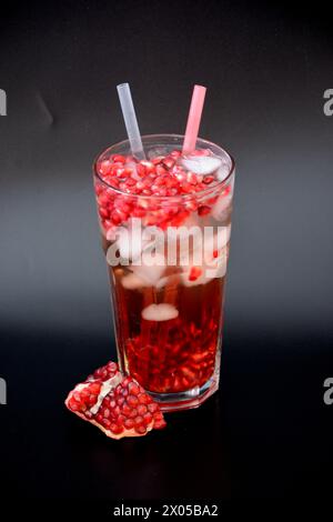 Jus de grenade rafraîchissant avec de la glace et des graines dans un grand verre sur fond noir, à côté d'un fruit cassé. Disposition verticale. Banque D'Images