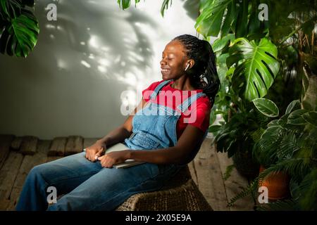 Joyeuse jeune femme noire porter des écouteurs sourires se trouve au jardin de la maison, tient un ordinateur portable fermé sur les genoux Banque D'Images