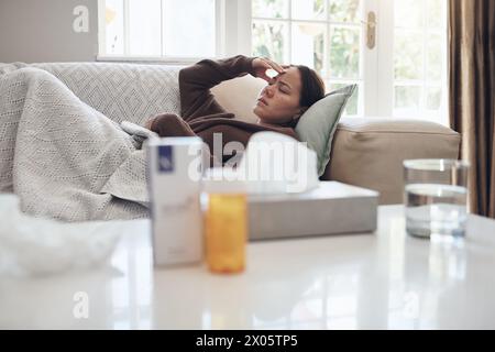 Femme fatiguée, malade et mal de tête avec des médicaments sur le canapé pour la grippe, le rhume ou les sinus dans le salon à la maison. Jeune femme avec maladie, fièvre ou Banque D'Images