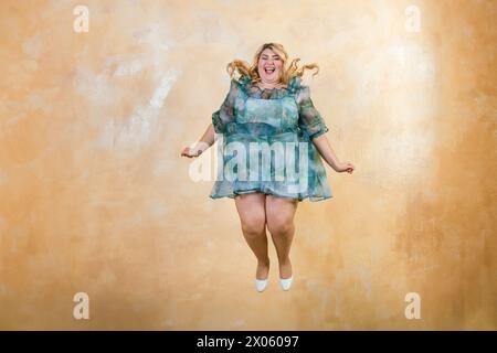 Excité femme plus taille dans la robe verte sautant dans la joie sur fond jaune dans le studio Banque D'Images
